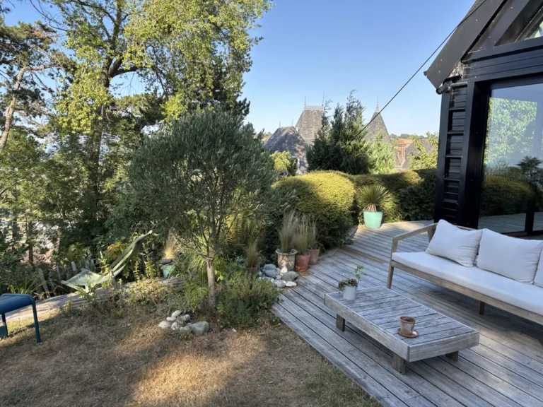 Holzterrasse mit weißem Sofa, Couchtisch und Olivenbaum, spitze Dächer im Hintergrund