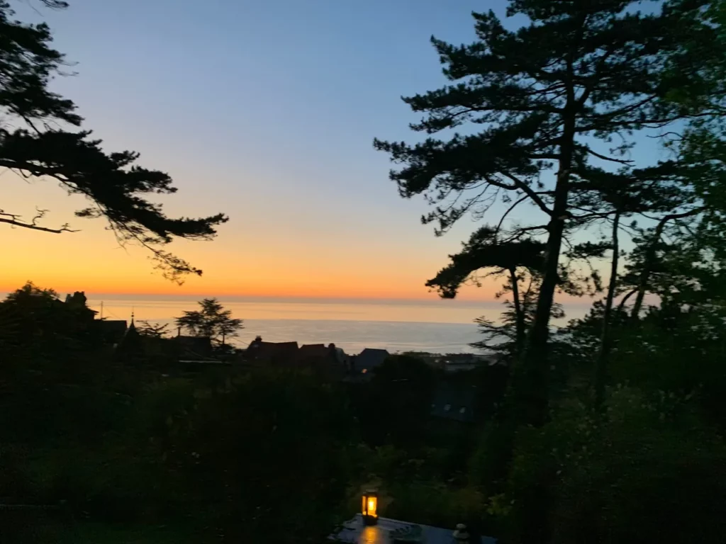 Orangefarbener Sonnenuntergang über dem Meer zwischen den Seekiefern, brennende Laterne im Vordergrund, vom Garten des Carré Vue Mer in Pourville-sur-Mer
