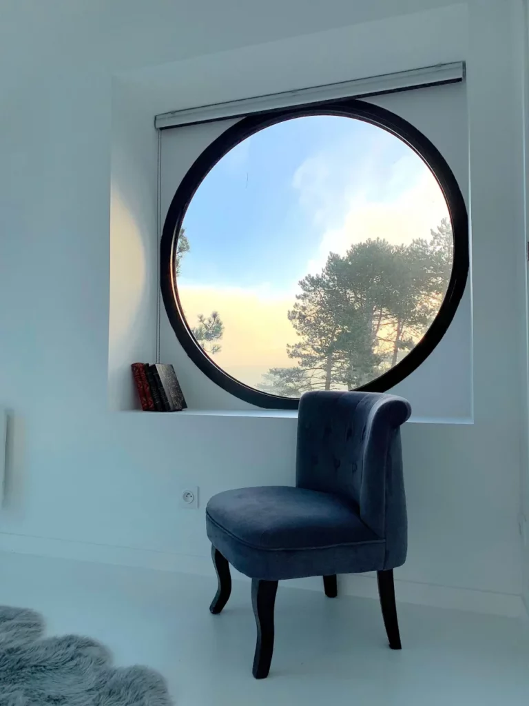 Rundes Bullauge und blauer Samtsessel im Zimmer Est des Carré Vue Mer, Kiefern und Himmel bei Sonnenaufgang