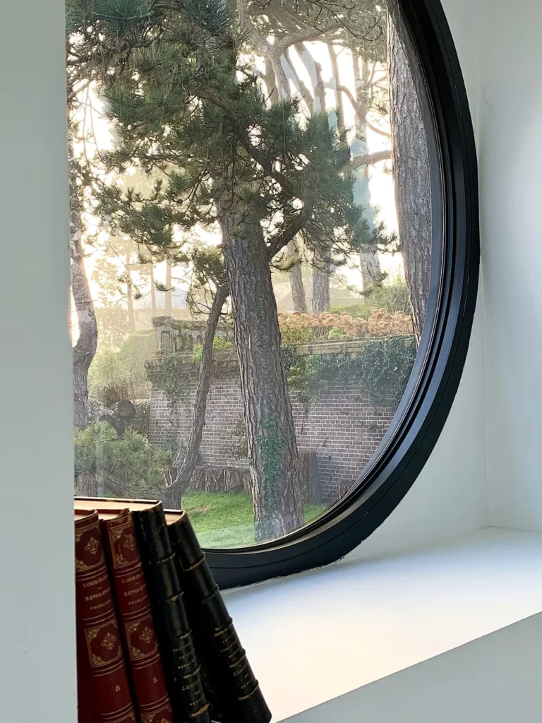 Rundes Bullauge mit Blick auf die Seekiefern und eine alte Backsteinmauer, alte gebundene Bücher auf der Fensterbank, Villa Le Carré Vue Mer in Pourville
