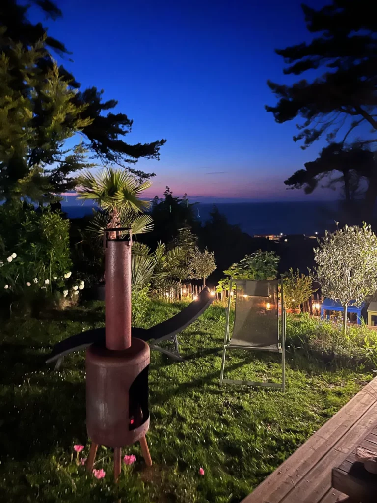 Der Garten des Carré Vue Mer bei Einbruch der Nacht: Feuerschale, Liegestühle, Palme und Lichterketten, das Meer in der Ferne in Pourville-sur-Mer