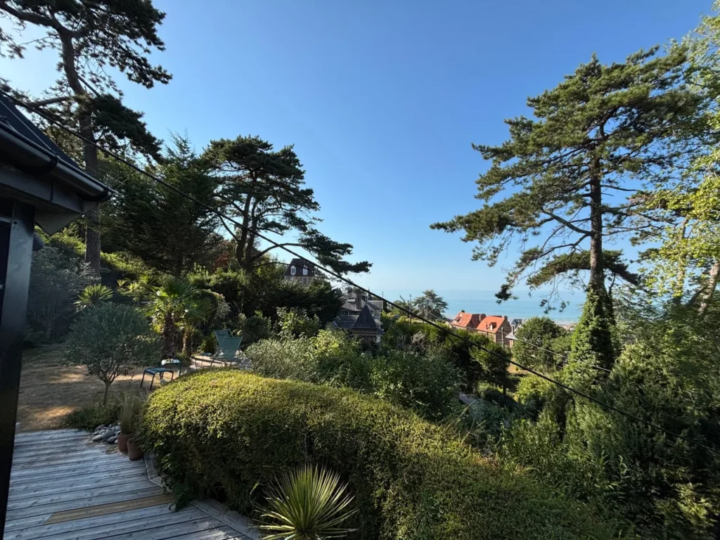 Terrassengarten unter hohen Seekiefern von der Holzterrasse, blaues Meer und Dächer von Pourville-sur-Mer im Sommer