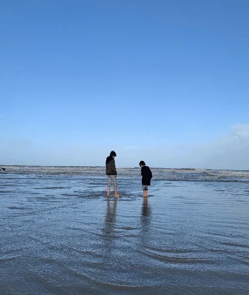 Zwei Kinder mit den Füßen im Wasser am Strand bei Ebbe unter blauem Himmel