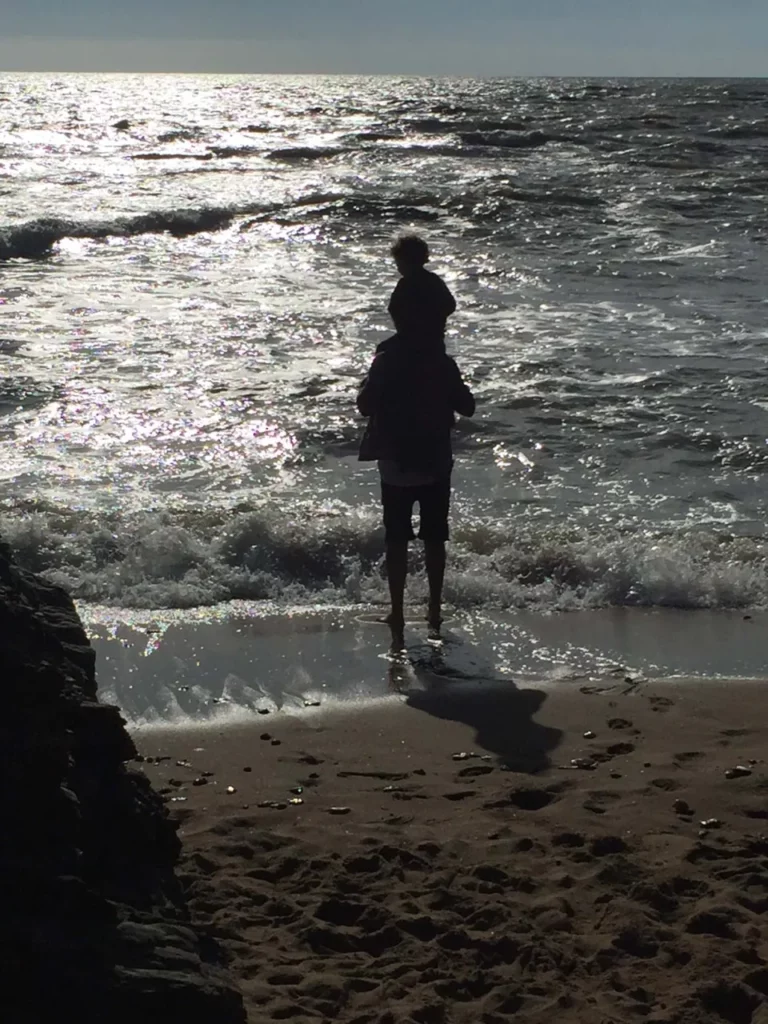 Gegenlicht-Silhouette eines Kindes auf den Schultern eines Erwachsenen am Meer