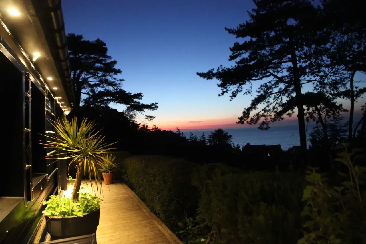 Terrasse en bois éclairée à la tombée de la nuit, palmier et pins, coucher de soleil sur la mer à Pourville-sur-Mer