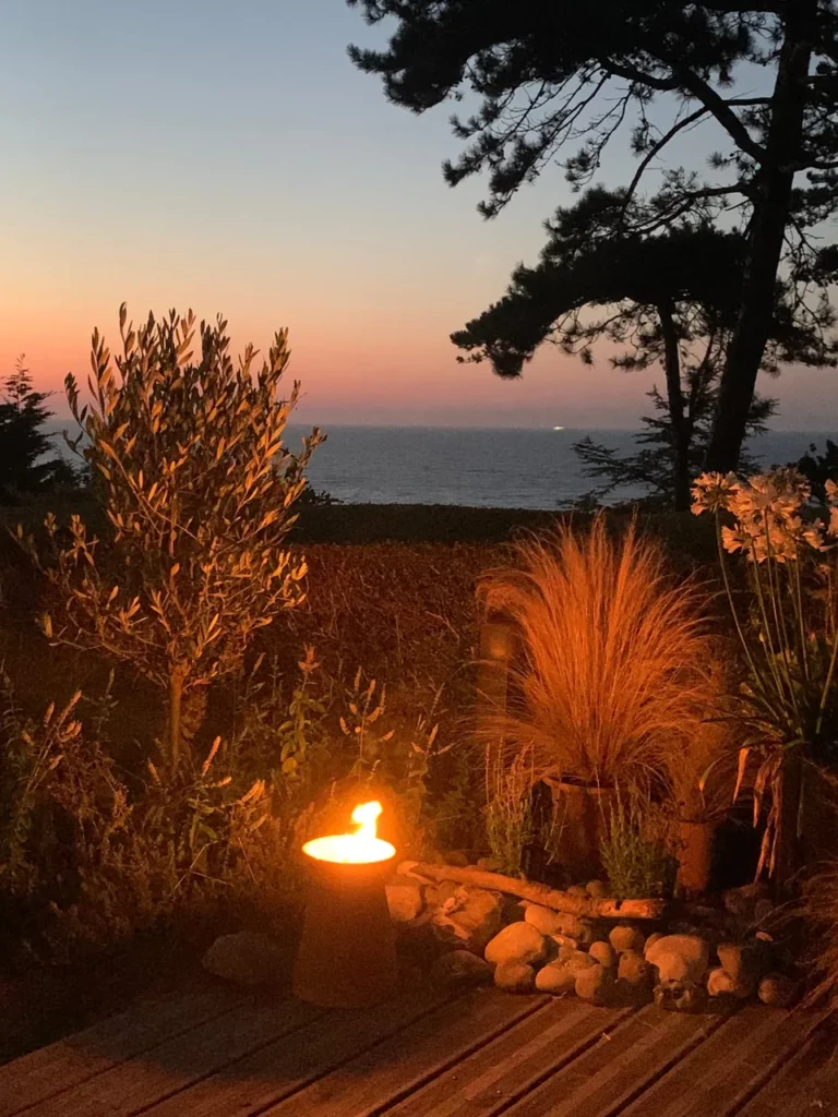 Brennende Fackel auf der Holzterrasse bei Sonnenuntergang, Olivenbaum, Gräser und Schmucklilien, Meer und Seekiefer in der Ferne in Pourville-sur-Mer
