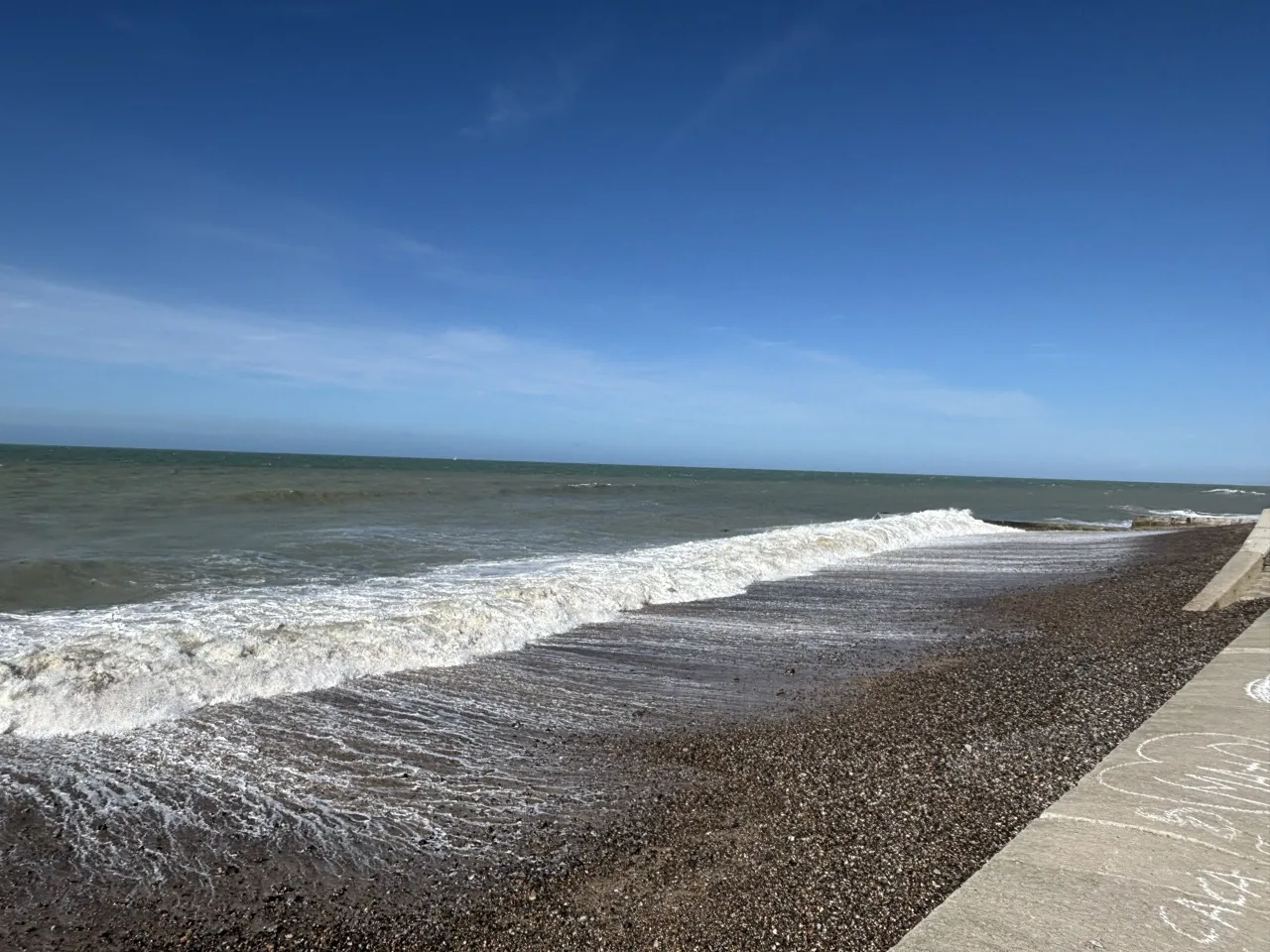 Vague déferlant sur la plage de galets de Pourville-sur-Mer sous un ciel bleu