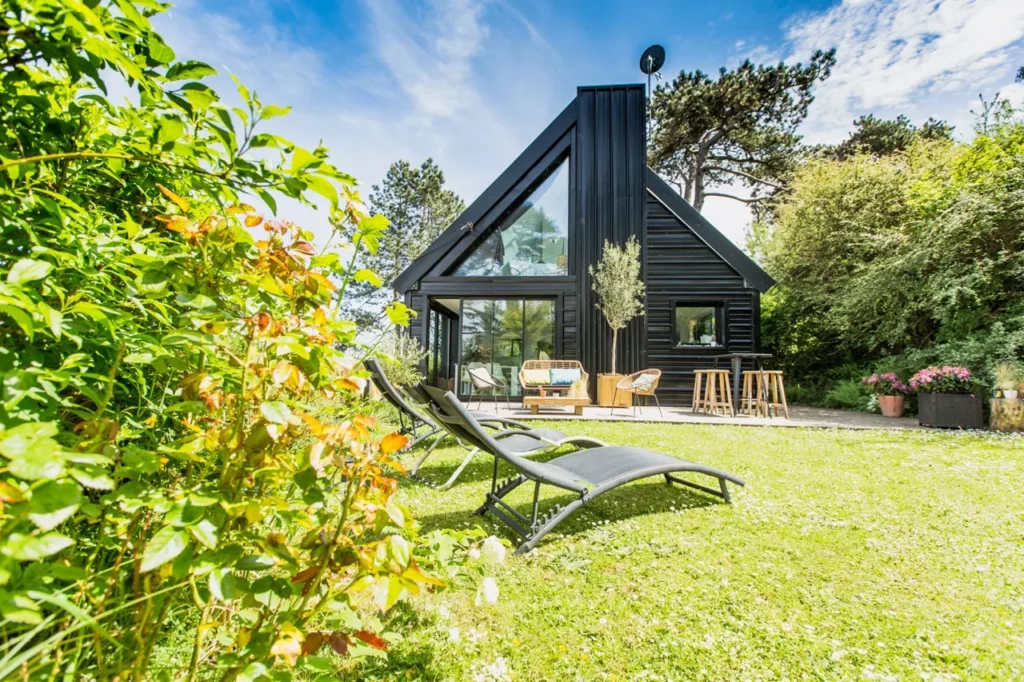 Villa en bois noir avec grande verrière, terrasse et transats sur la pelouse ensoleillée